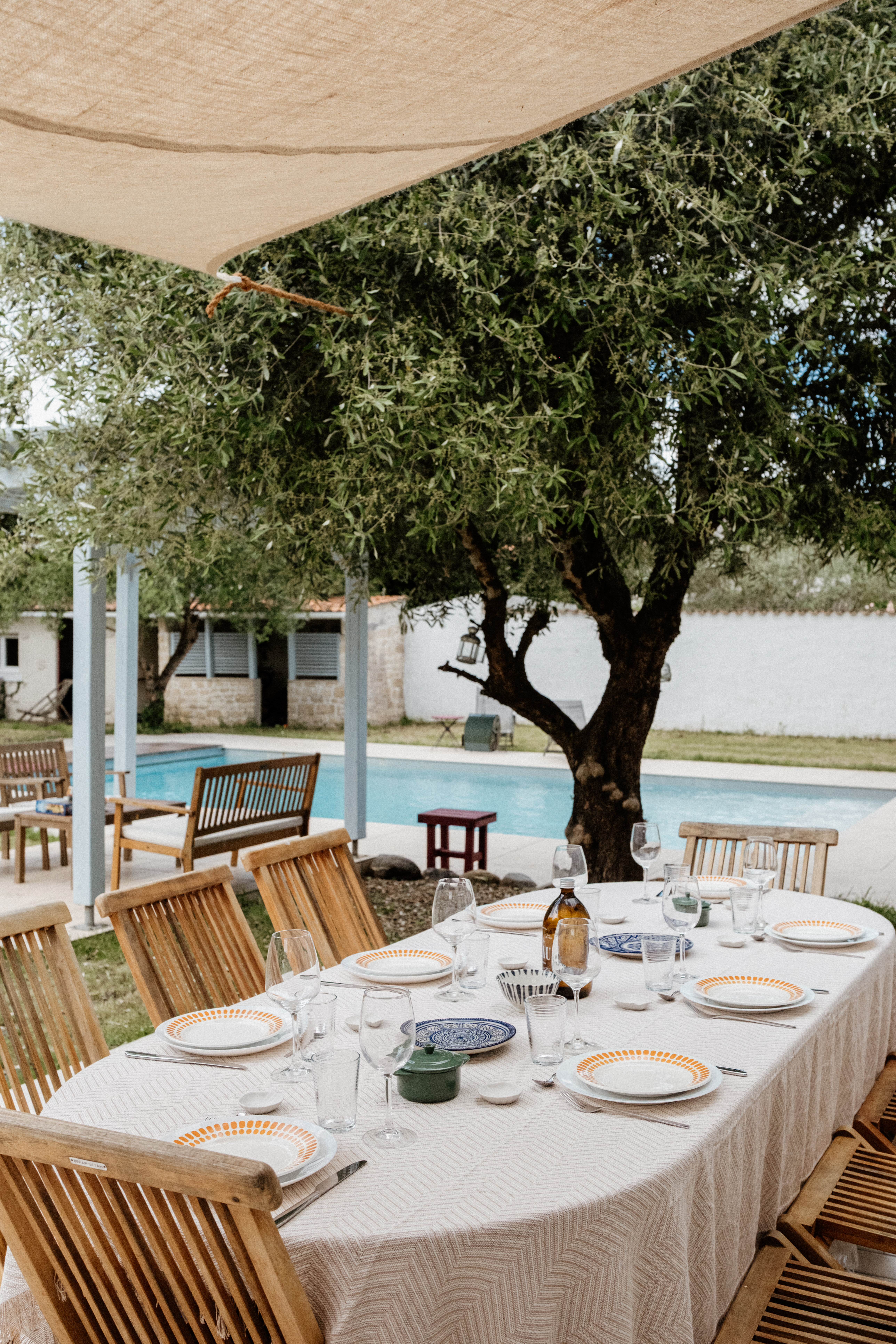 Long outdoor table set for a meal under a shade sail beside an olive tree, with a pool and stone house behind
