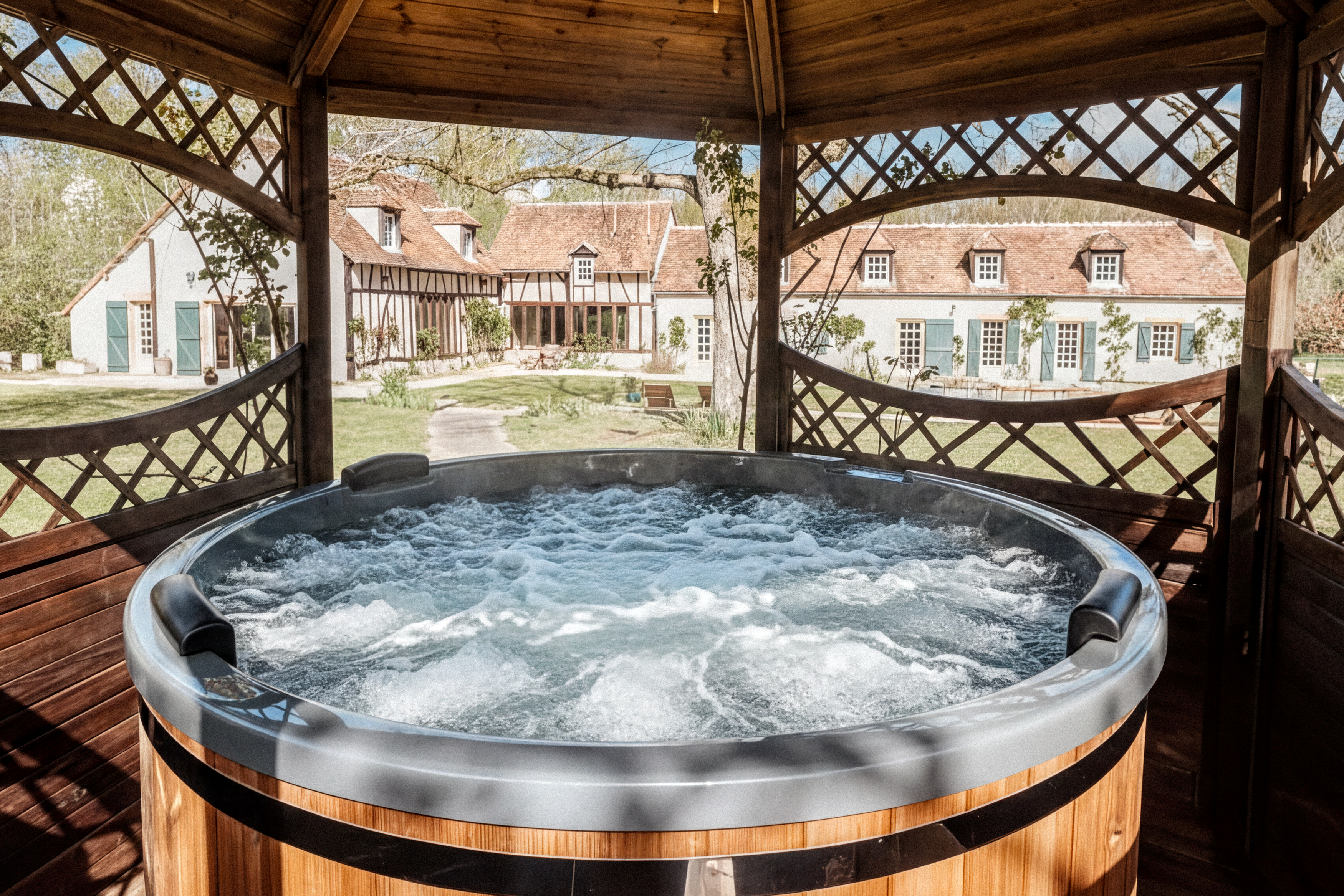 Cedar hot tub framed through a gazebo lattice with a half-timbered Normandy farmhouse beyond